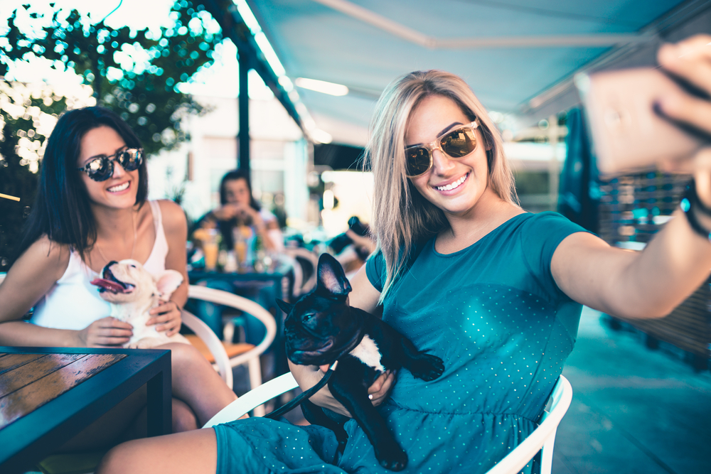 Two women wearing sunglasses, one holding a small black dog, taking a selfie at an outdoor cafe. They are smiling and dressed in casual summer attire, after visiting veterinary care in Houston.