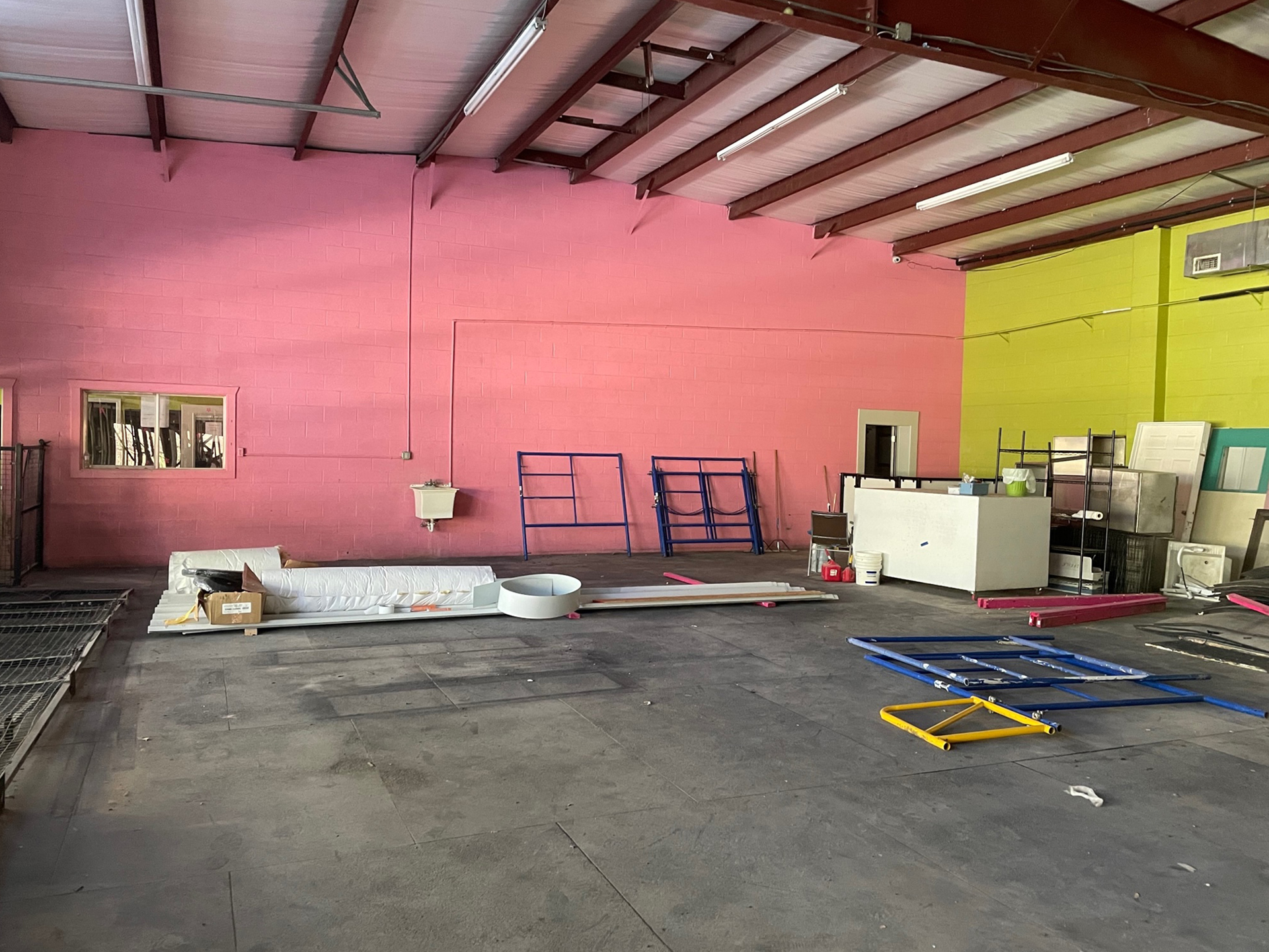 Interior of a spacious industrial warehouse with pink and green walls, featuring scattered equipment like ladders and containers, and a mattress on the floor for pet rehabilitation.