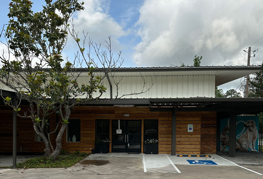 A modern single-story building with a wooden façade and extended white metal roof, featuring two handicap parking spaces in front under a partly cloudy sky, offering specialized services in pet behavior guidance.