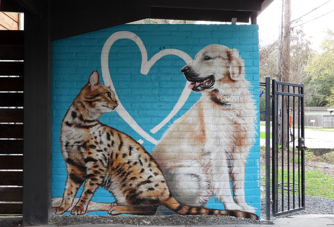 A mural of a golden retriever and a lynx sitting closely, both facing forward with a large white heart outline in the background, painted on a blue wall under a covered area dedicated to pet behavior