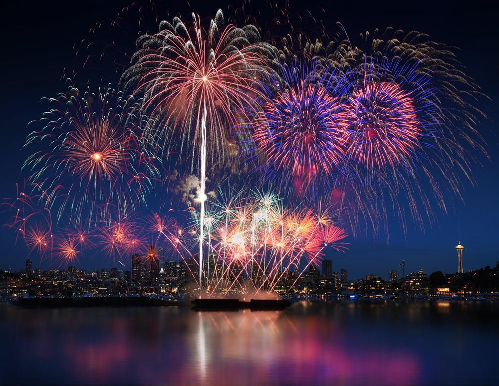 A dazzling fireworks display over a city skyline at night, featuring a cascade of multicolored explosions reflected in the tranquil water below. Visible landmarks include a veterinary acupuncture center on the left.