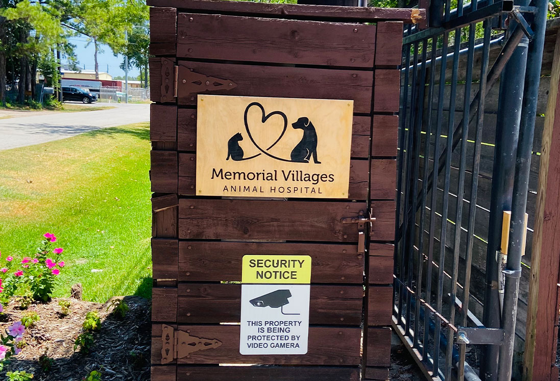 A wooden sign for Memorial Villages Animal Hospital features a logo with two pets and a heart, mounted on a fence with a security notice about video surveillance. Flowers are visible at the base. The sign