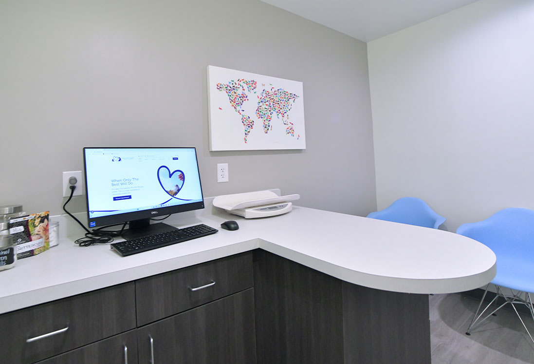 A modern veterinary medical office with a curved reception desk, computer displaying a heart health website, and two blue chairs. A world map artwork is on the wall.