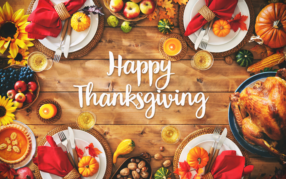 A festive Thanksgiving table setting featuring plates with autumn-themed napkins, small pumpkins, sunflowers, and a roasted turkey centerpiece. The text "Happy Thanksgiving" is written in the center on a wooden background.