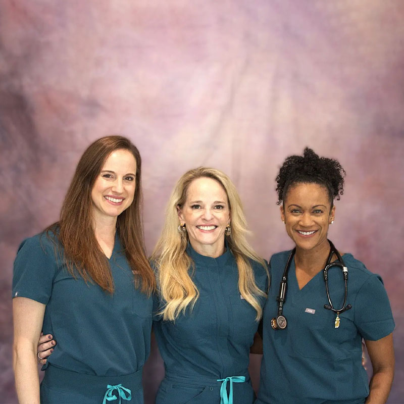 Three women wearing teal medical scrubs, one with a stethoscope around her neck, stand close together smiling in front of a blurred, light purple background.